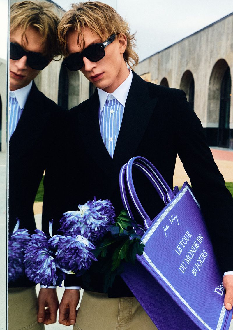 Model Aleksander Lukowski wearing black sunglasses, a black blazer, and holding a purple tote bag next to a mirror reflection.
