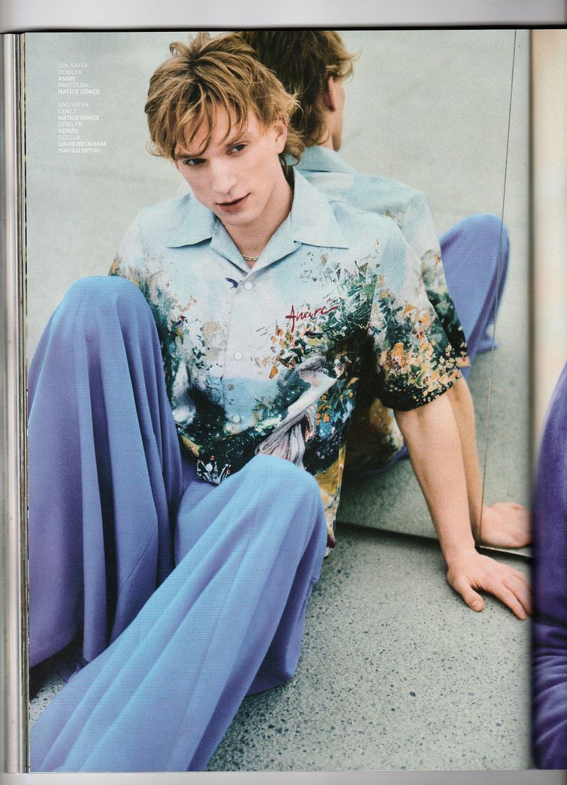 A blonde male model poses while seated in a button-down shirt and wide-legged pants, reflecting in a large mirror in a fashion editorial.