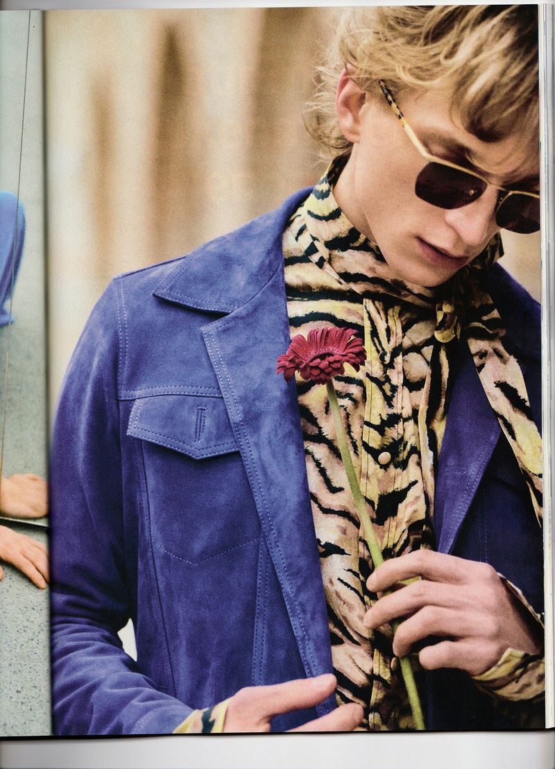 Model Aleksander Lukowski wearing a purple suede jacket, tiger print top, and sunglasses, holding a single red flower.