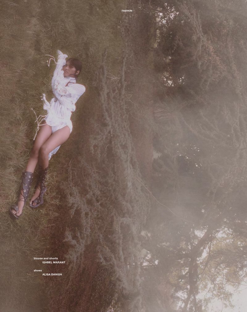 Model Ashley Radjarame lying on a grassy slope wearing a white textured blouse and knee-high lace-up sandals in hazy natural light.