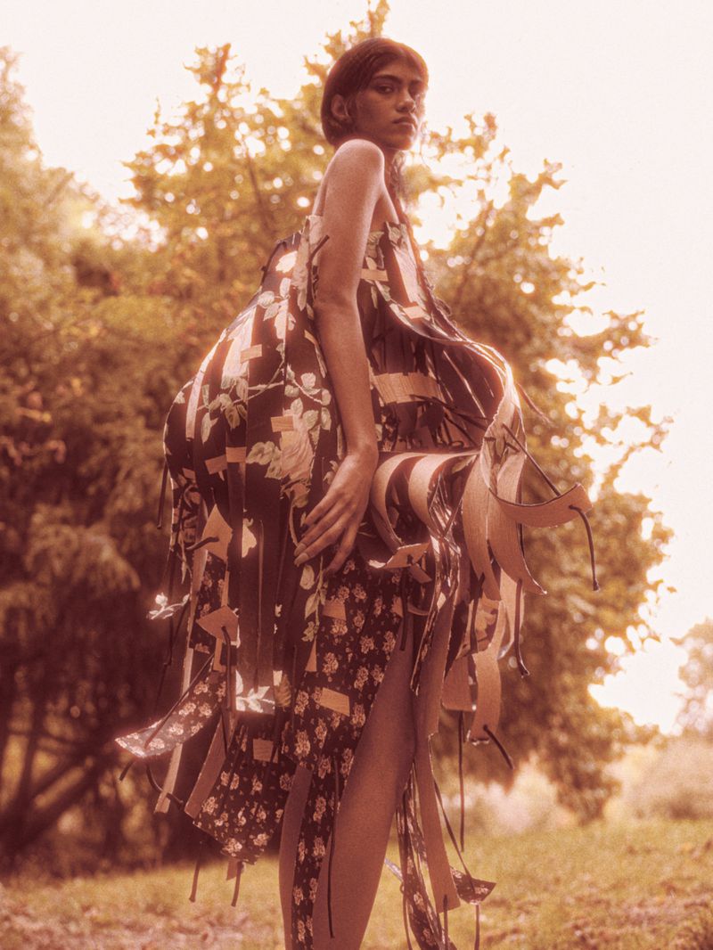 Model Ashley Radjarame standing outdoors in an avant-garde floral ribbon dress surrounded by trees in warm sunlight.
