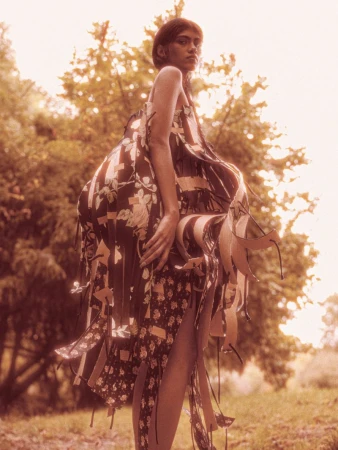 Model Ashley Radjarame standing outdoors in an avant-garde floral ribbon dress surrounded by trees in warm sunlight.