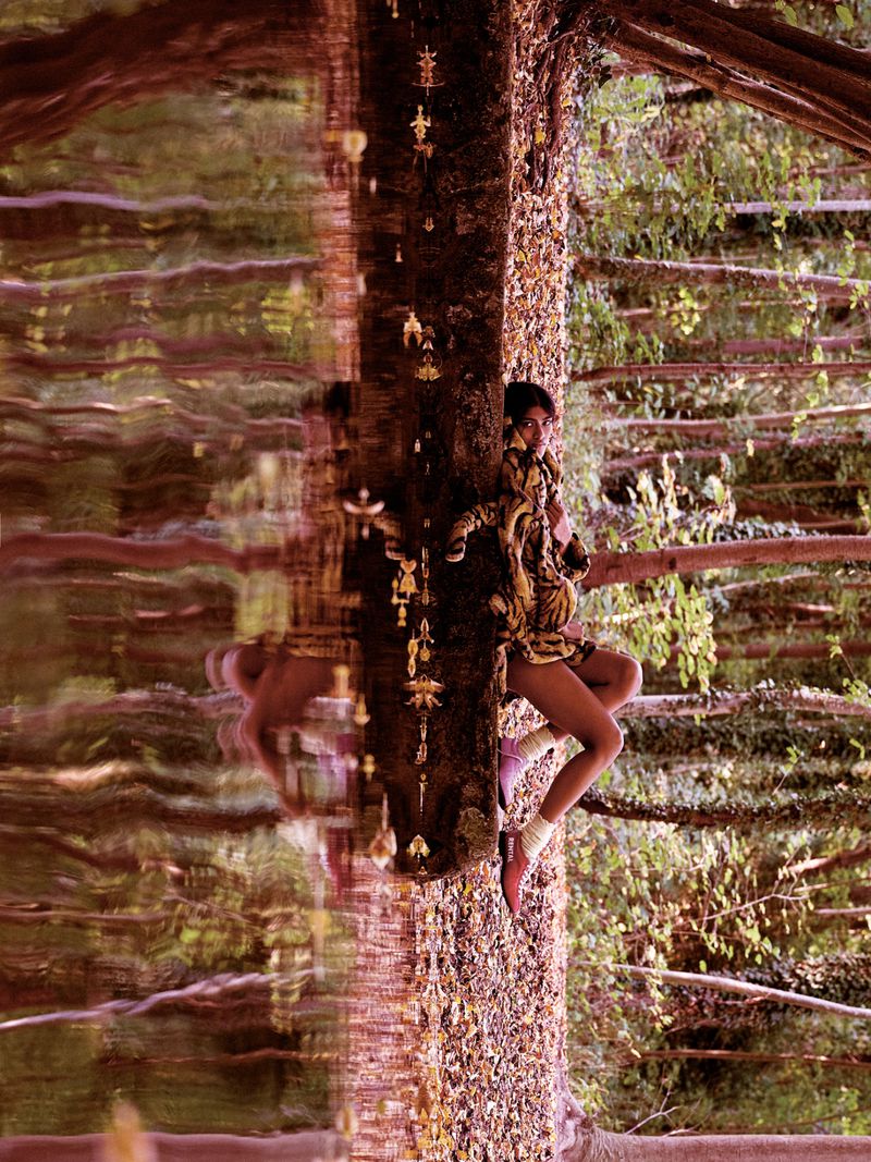 Model Ashley Radjarame in a patterned jacket and socks posing against a tree trunk reflected in rippling water in a forest.