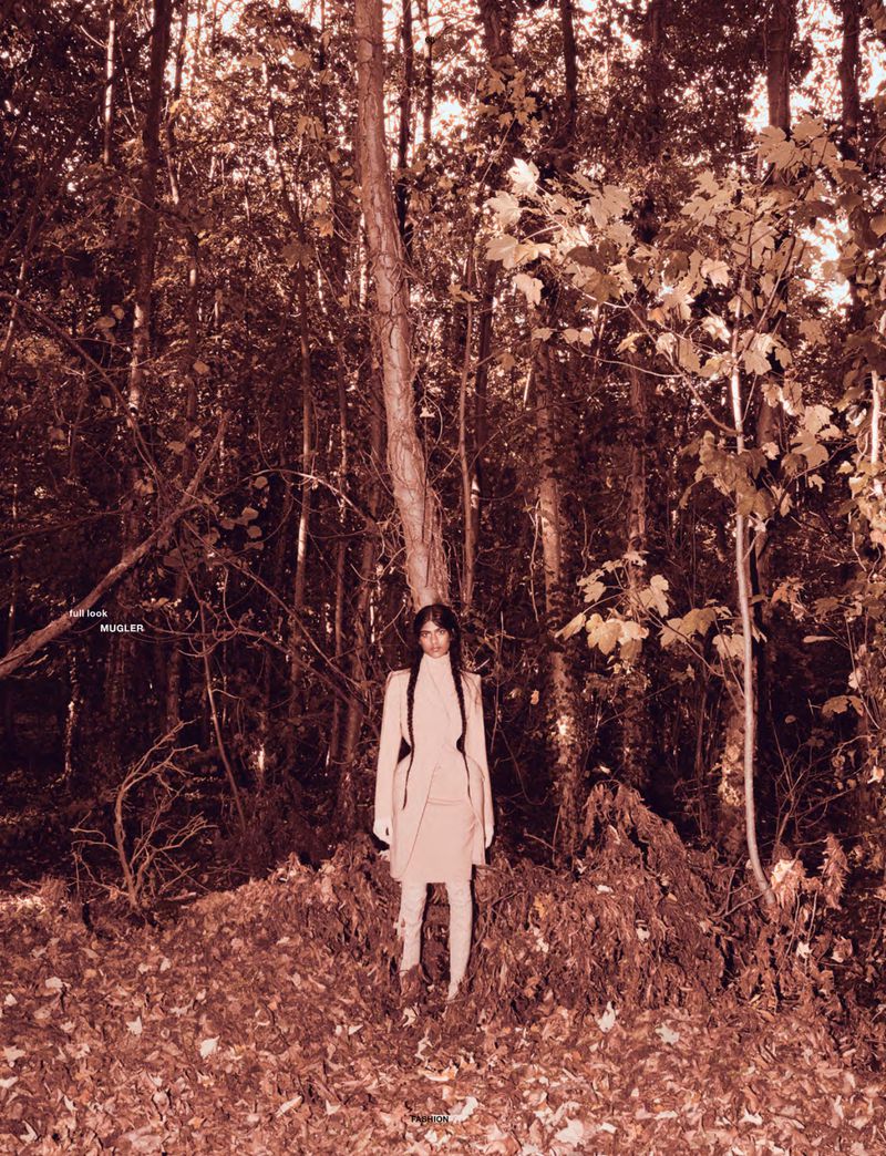 Model Ashley Radjarame standing in a tailored light beige coat amidst dense trees and fallen autumn leaves in a forest.