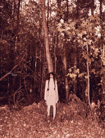 Model Ashley Radjarame standing in a tailored light beige coat amidst dense trees and fallen autumn leaves in a forest.