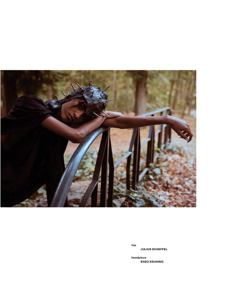 Model Ashley Radjarame wearing a dark spiked headpiece leans across a metal bridge railing outdoors in a woodland setting.