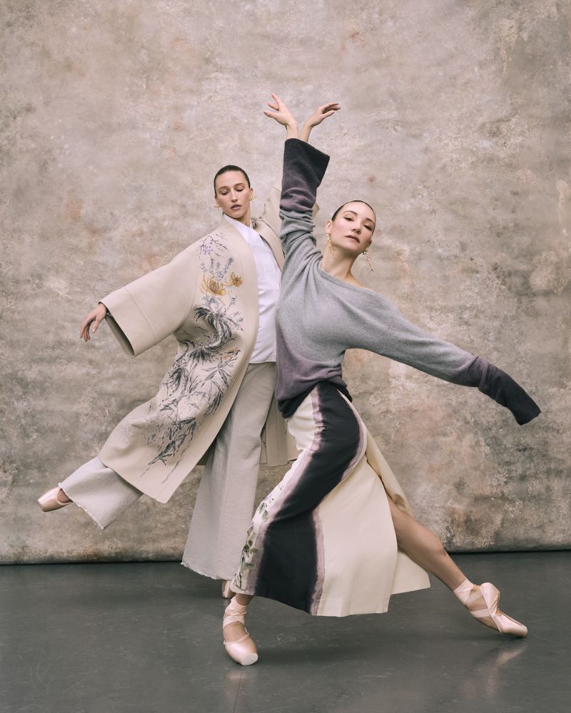Two ballet dancers in studio. One wears a long beige coat with floral detail, the other wears a grey sweater and white skirt.