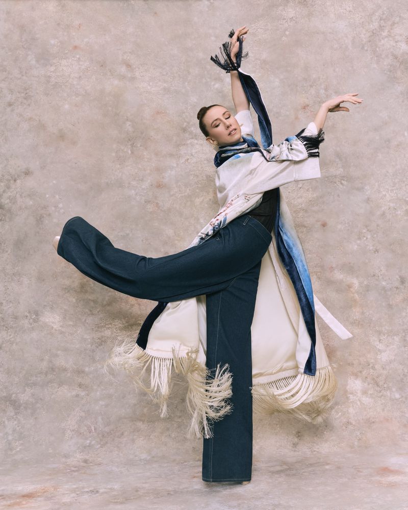 A slender woman strikes a dynamic dance pose in wide-leg jeans and a fringed white robe against a mottled backdrop, showcasing fashion and movement.