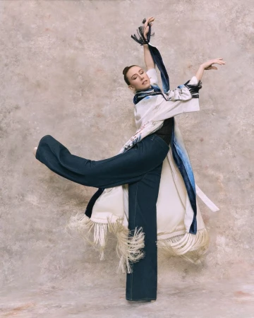 A slender woman strikes a dynamic dance pose in wide-leg jeans and a fringed white robe against a mottled backdrop, showcasing fashion and movement.