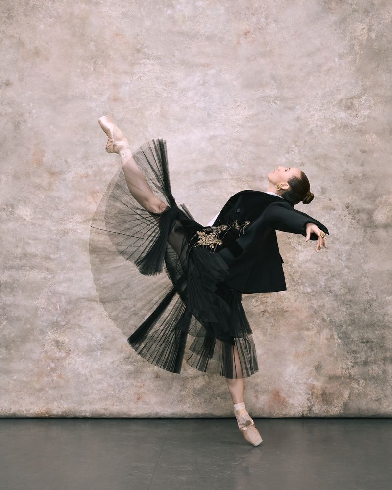 Elegant adult woman performing ballet in black skirt and blazer against a textured neutral background. Dance and high fashion concept.