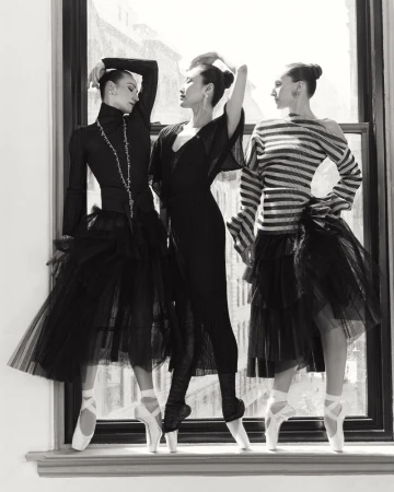Three ballerinas in pointe shoes and tulle skirts pose in front of a bright window, dressed in black and white, shot in black and white.