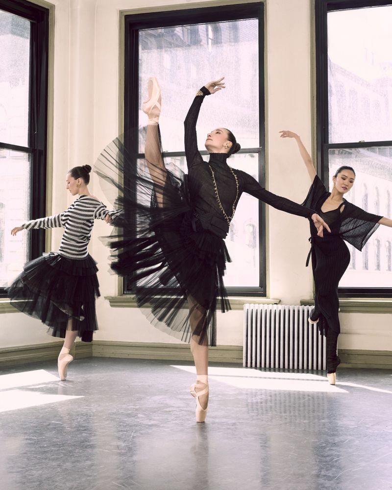 Three ballerinas in black attire gracefully pose in a dance studio with ballet shoes and tutus, lit by natural light streaming through windows.