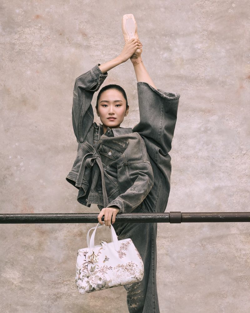 Woman in denim outfit with ballet shoes does a leg split at barre, holding a patterned white handbag in studio setting.