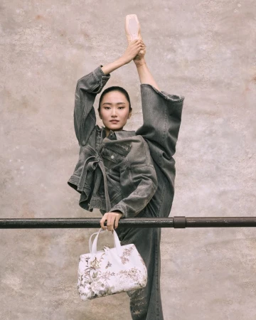 Woman in denim outfit with ballet shoes does a leg split at barre, holding a patterned white handbag in studio setting.