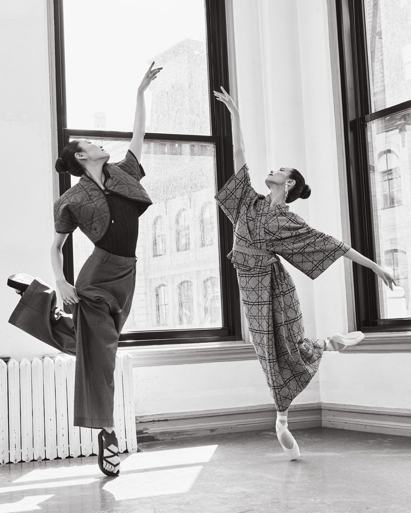 Two ballerinas en pointe pose in front of a large window in a dance studio. One wears an oversized kimono and the other an avant-garde outfit.