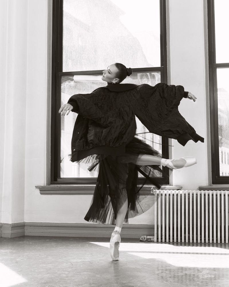 Monochrome image of a female ballet dancer in a black tutu and jacket, en pointe, with a window and radiator in the background.