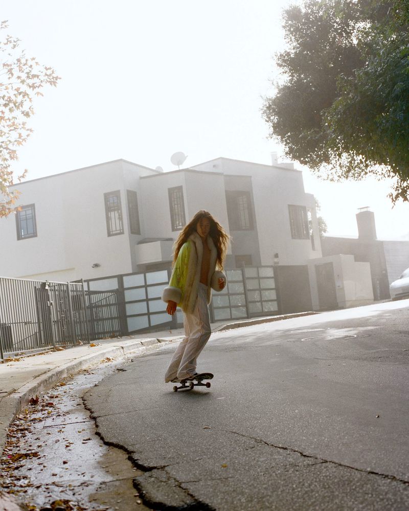 Model Elan Lee rides a skateboard down a sunlit residential street wearing a shearling-trimmed jacket and light trousers.