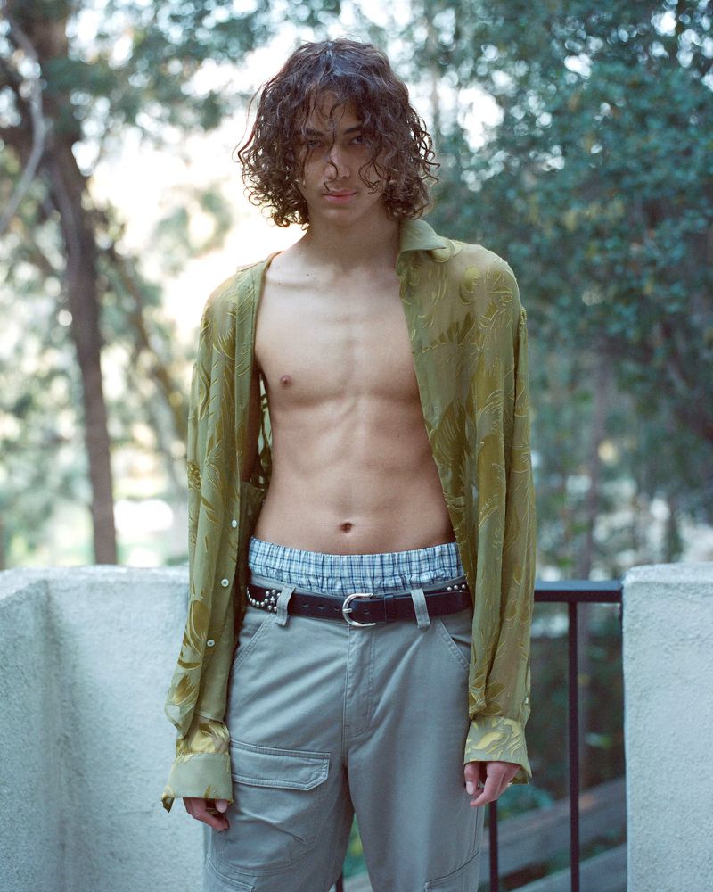 Model Elan Lee standing outdoors wearing an open sheer olive-green patterned shirt and grey cargo pants on a balcony.