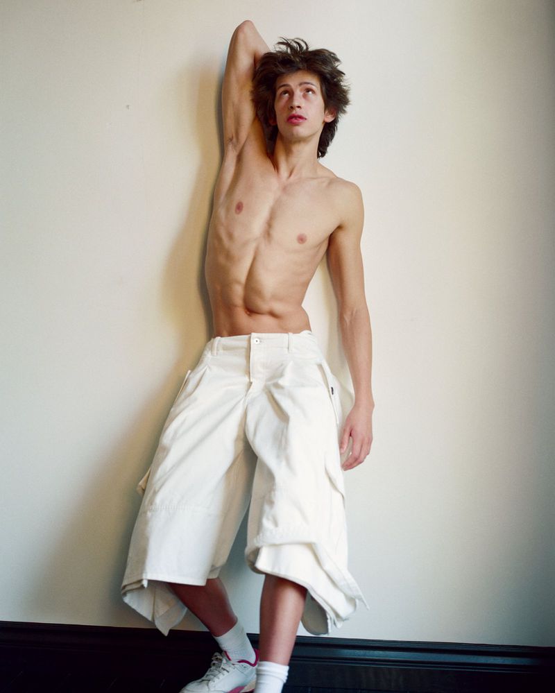 Shirtless male model Elan Lee wearing oversized white shorts leaning against a plain wall for Arena Homme+.