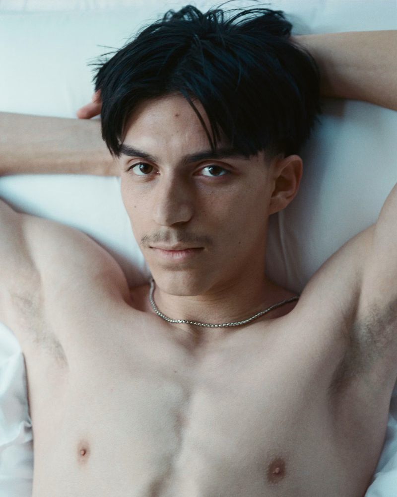 Portrait of a shirtless young adult male with dark hair, lying on a white sheet with his hands behind his head, looking at the camera.