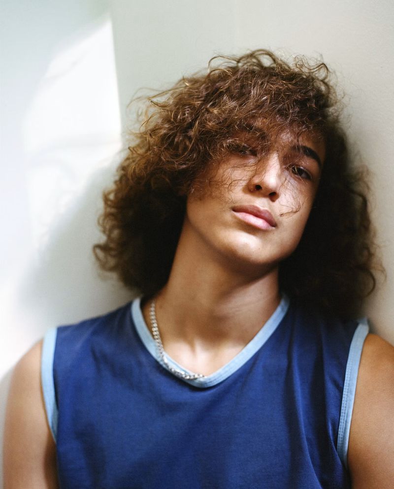 Model Elan Lee with curly brown hair wearing a blue sleeveless shirt leaning against a pale wall in a close-up portrait.