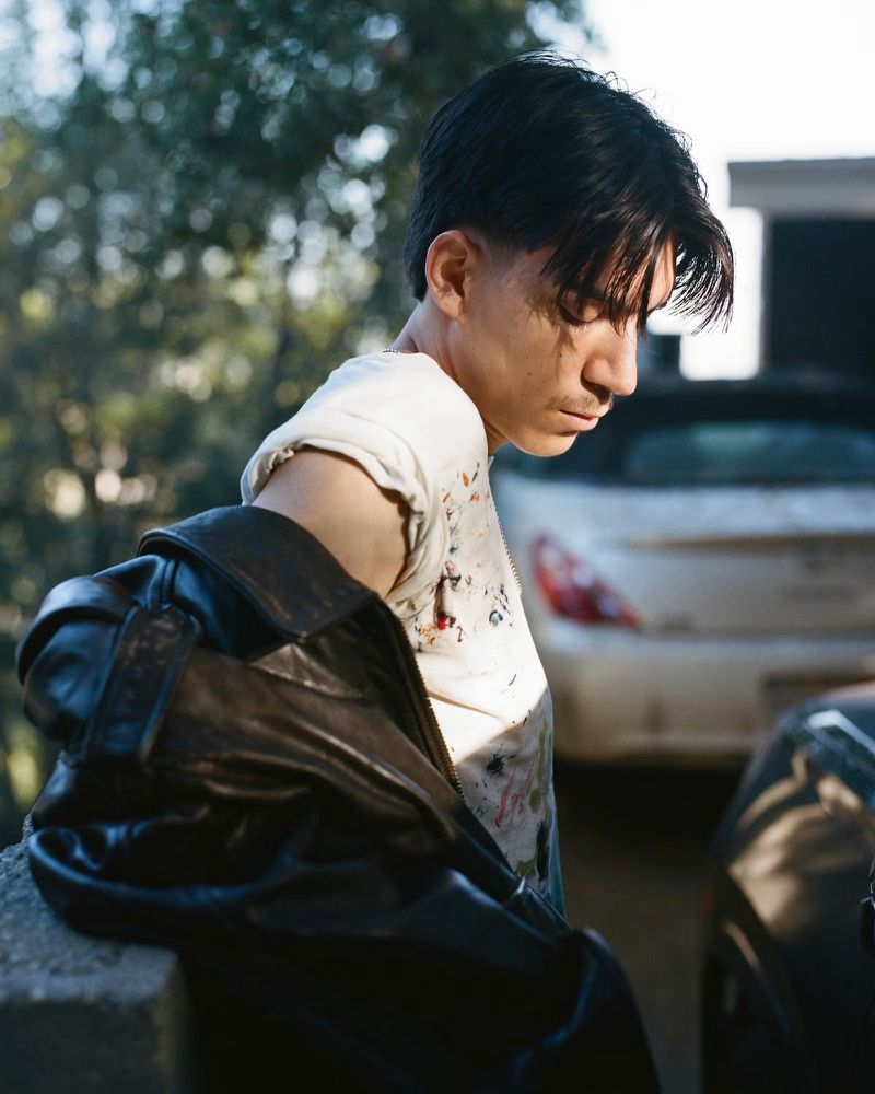 Model Elan Lee looking down while holding a black leather jacket and wearing a white t-shirt outdoors in sunlight.