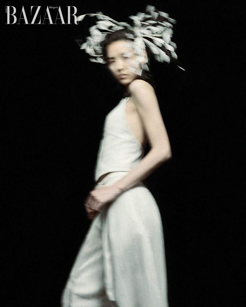 Model Song Ah Woo wearing a cream halter top and feathered headpiece with motion blur against a solid black studio background.