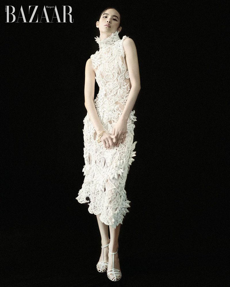 Model Song Ah Woo wearing a textured white lace high-neck dress and strappy sandals against a black backdrop for Harper's Bazaar Korea.