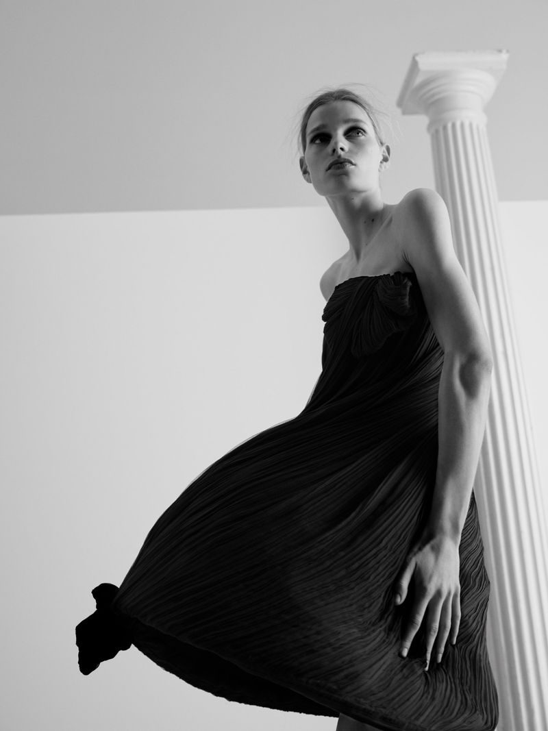 Saar Mansvelt Beck wearing a black pleated strapless dress posing beside a white fluted column in a low-angle shot.