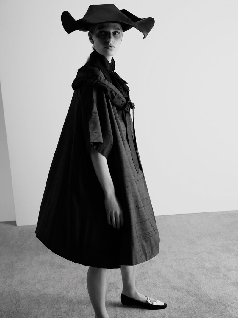 Model in wide-brimmed black hat and dark sculptural dress standing against a plain backdrop for Beyond Noise