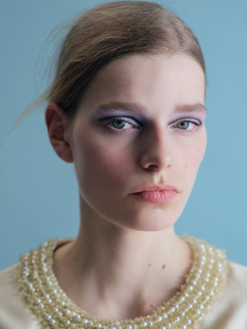 Close-up portrait of model Saar Mansvelt Beck wearing purple eye makeup and an embellished collar against a blue background