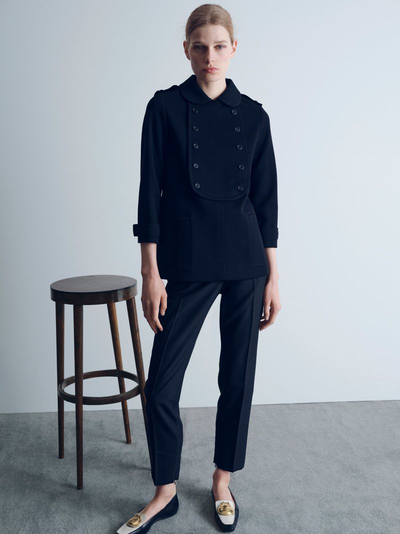 Model Saar Mansvelt Beck standing in a black double-breasted jacket and trousers beside a wooden stool in a minimalist studio.