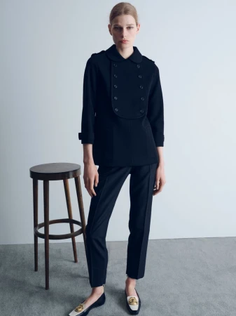 Model Saar Mansvelt Beck standing in a black double-breasted jacket and trousers beside a wooden stool in a minimalist studio.