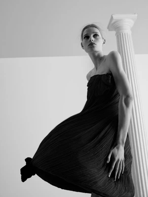 Saar Mansvelt Beck wearing a black pleated strapless dress posing beside a white fluted column in a low-angle shot.