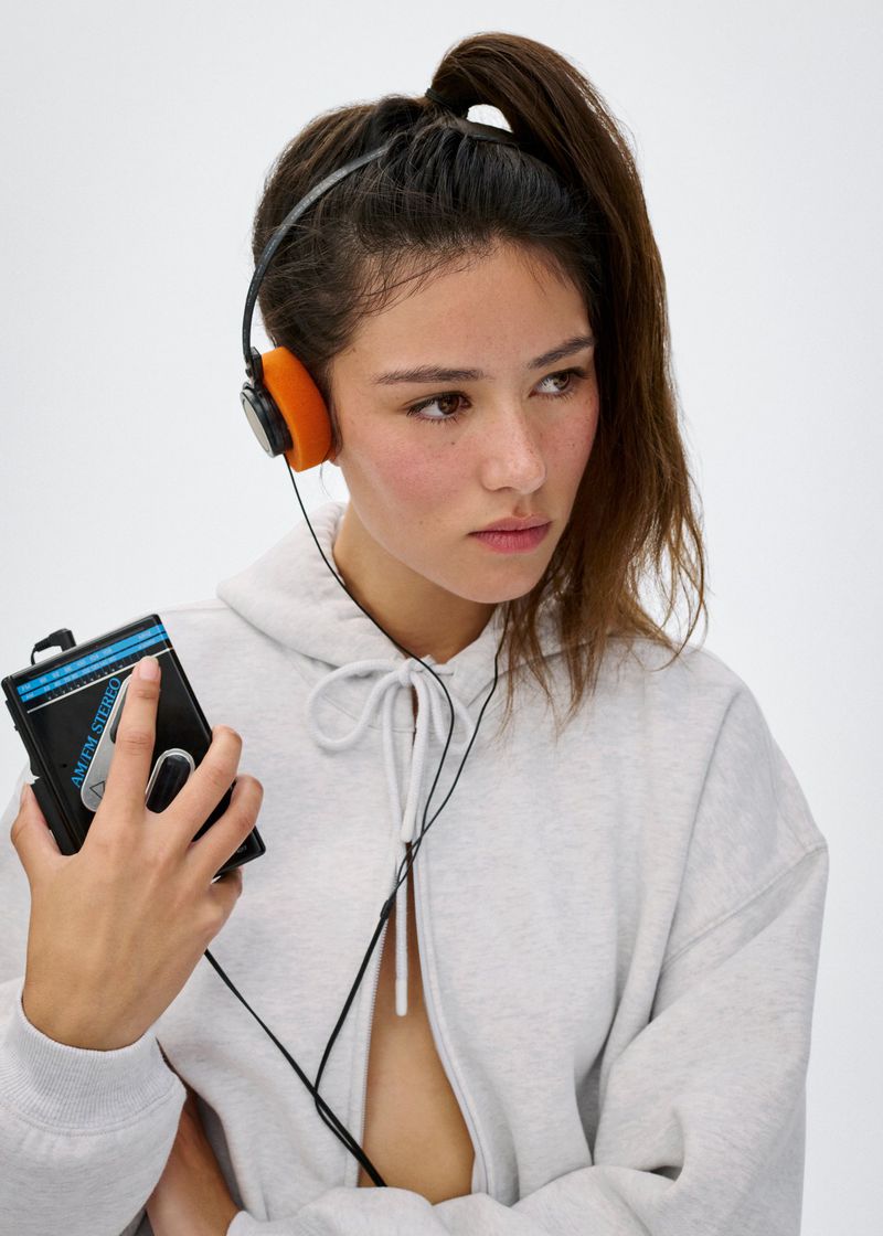 Model wearing a light grey hoodie and vintage headphones holding a portable cassette player against a white background.