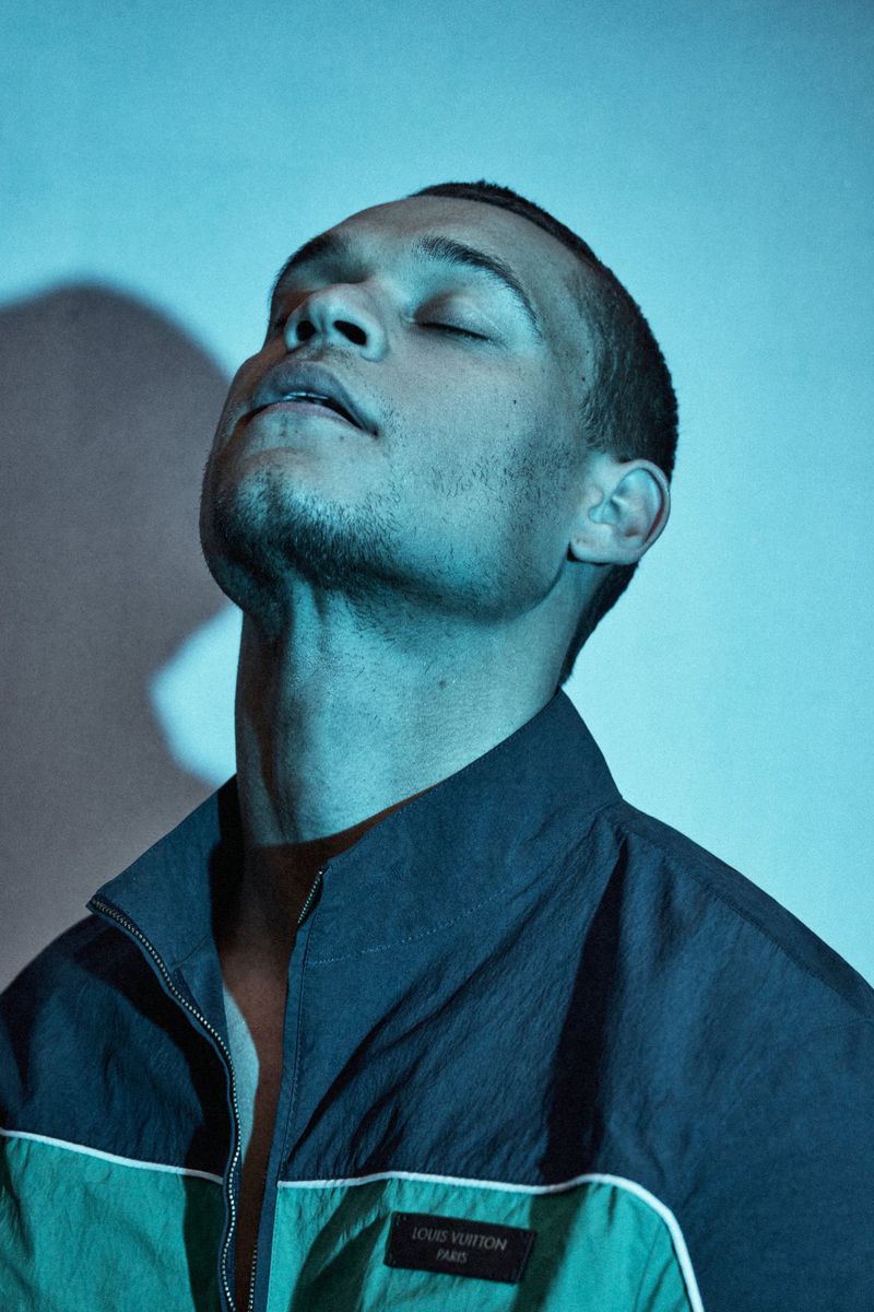 Tyriq Withers tilts his head back with eyes closed wearing a dark green and navy zip-up jacket against a blue-lit wall.