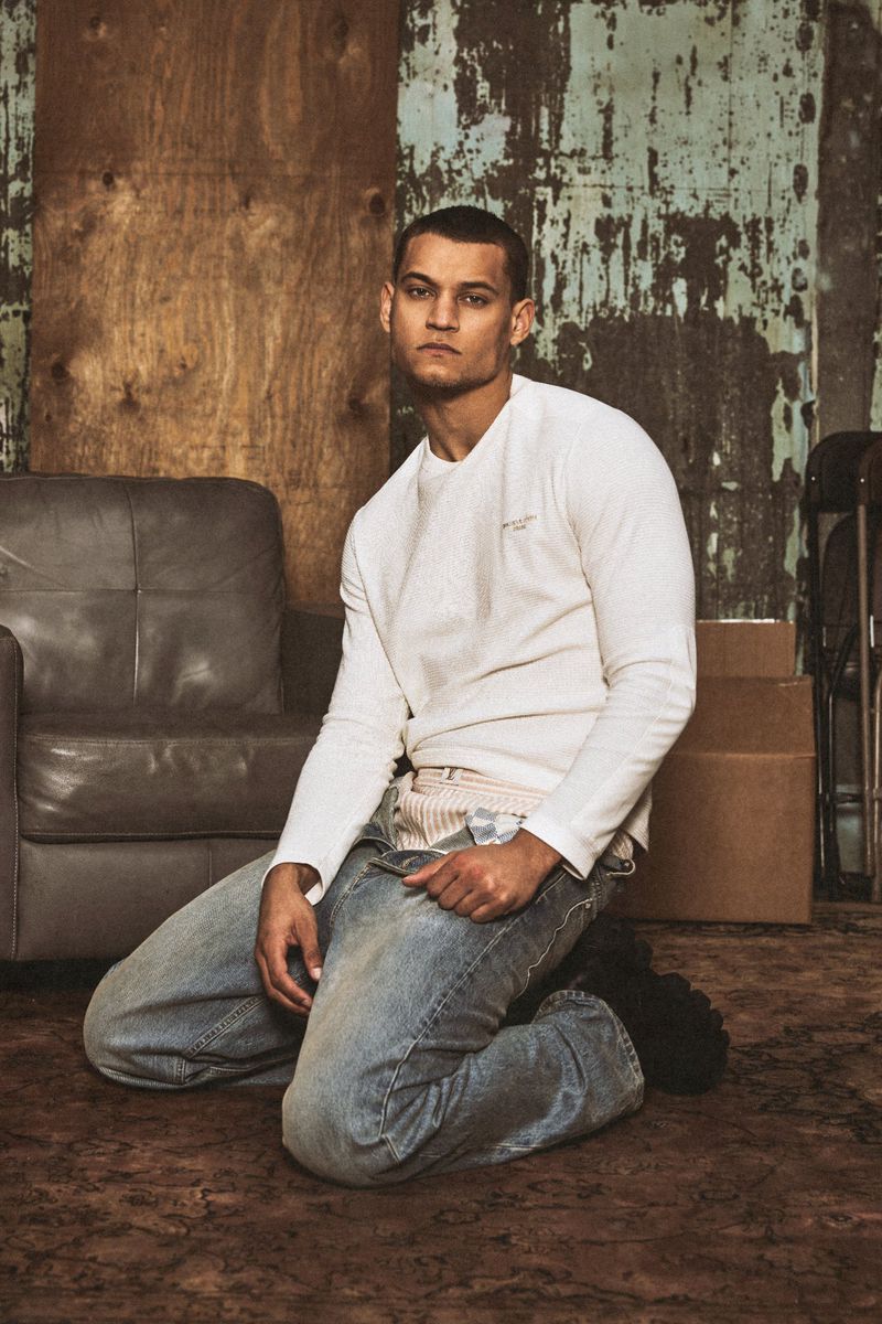 Actor Tyriq Withers kneeling in a white long-sleeve shirt and light blue jeans beside a grey armchair against a distressed wall.