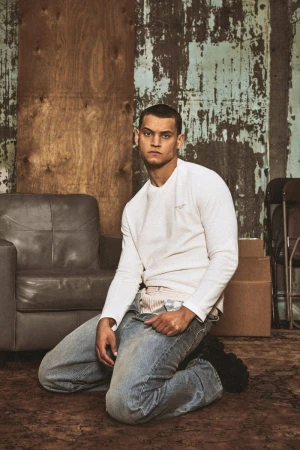 Actor Tyriq Withers kneeling on a rug wearing a white long-sleeve top and faded jeans in a set-dressed studio.