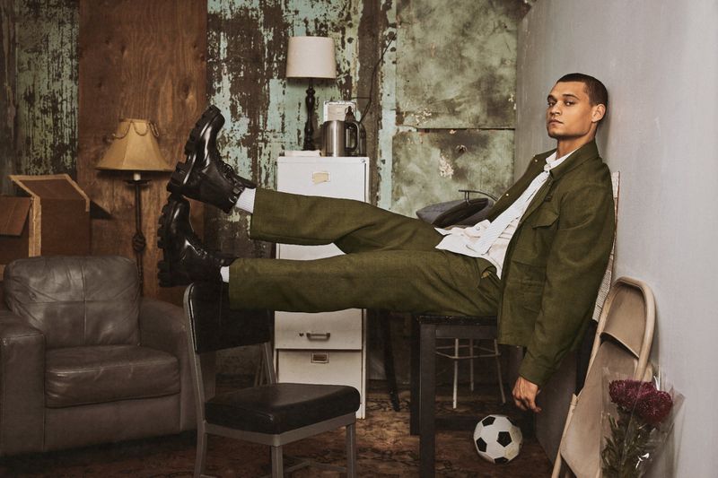 Tyriq Withers posing in an olive green suit and black chunky boots while reclining across furniture in a distressed room.