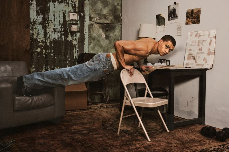 Shirtless actor Tyriq Withers performs an elevated pushup between an armchair and a folding chair in a rustic room.