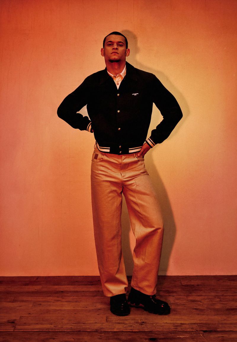 Tyriq Withers posing in a black jacket and tan wide-leg trousers against a warm studio wall with wooden floors.