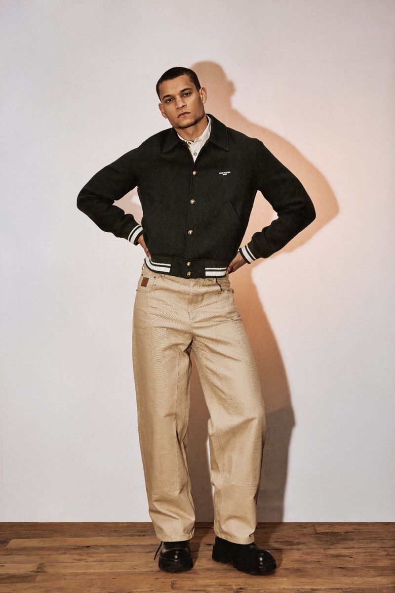 Full-length studio portrait of a man in a dark jacket, light patterned pants, and black boots on a wooden floor against a neutral wall.