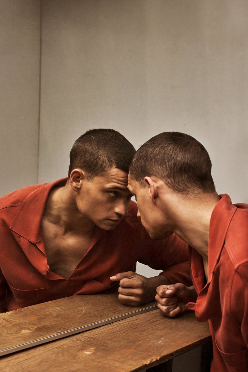 Actor Tyriq Withers in an open rust-colored shirt leans forward against a table mirror reflecting his face.