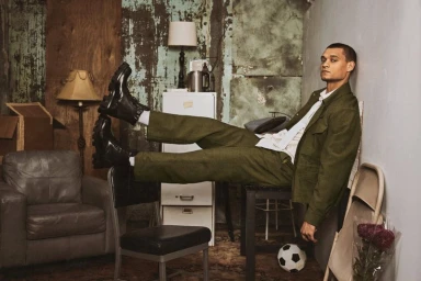 Tyriq Withers posing in an olive green suit and black chunky boots while reclining across furniture in a distressed room.