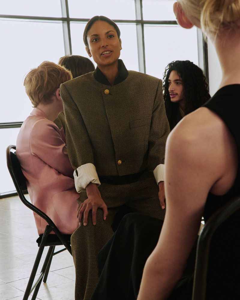 A seated model in an oversized olive houndstooth blazer speaks while surrounded by other models in front of large windows.