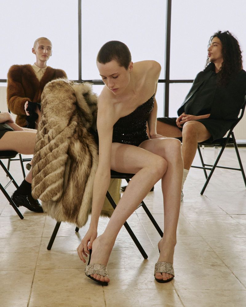 Model with a buzzcut in a black sequined top adjusting her mule sandal while seated in a chair beside a fur coat in a bright studio.
