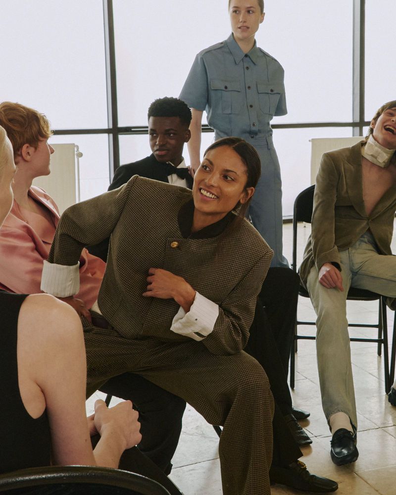 A smiling woman in a structured brown tailored suit sits among a group of fashion models in front of large windows.