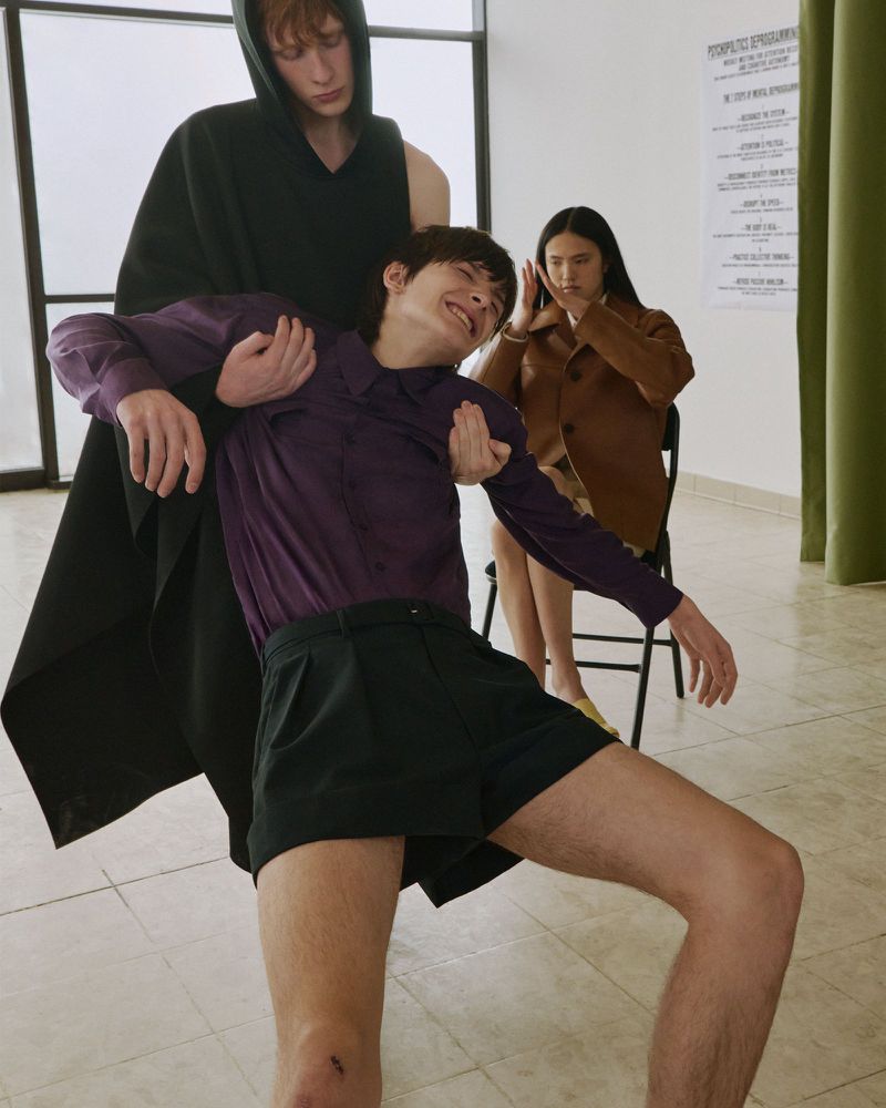 A model in a purple shirt and black shorts poses dynamically while supported by another model, with a seated woman in the background.