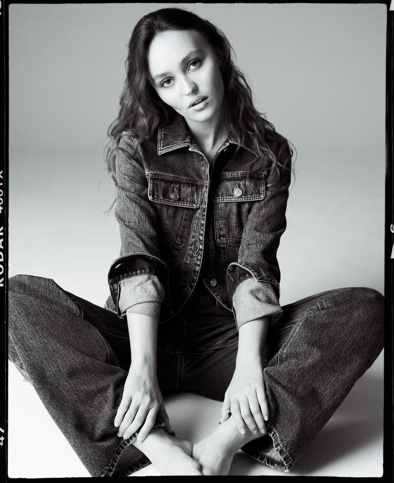 Lily-Rose Depp seated on the floor wearing a denim jacket and wide-leg jeans in a black-and-white studio portrait.
