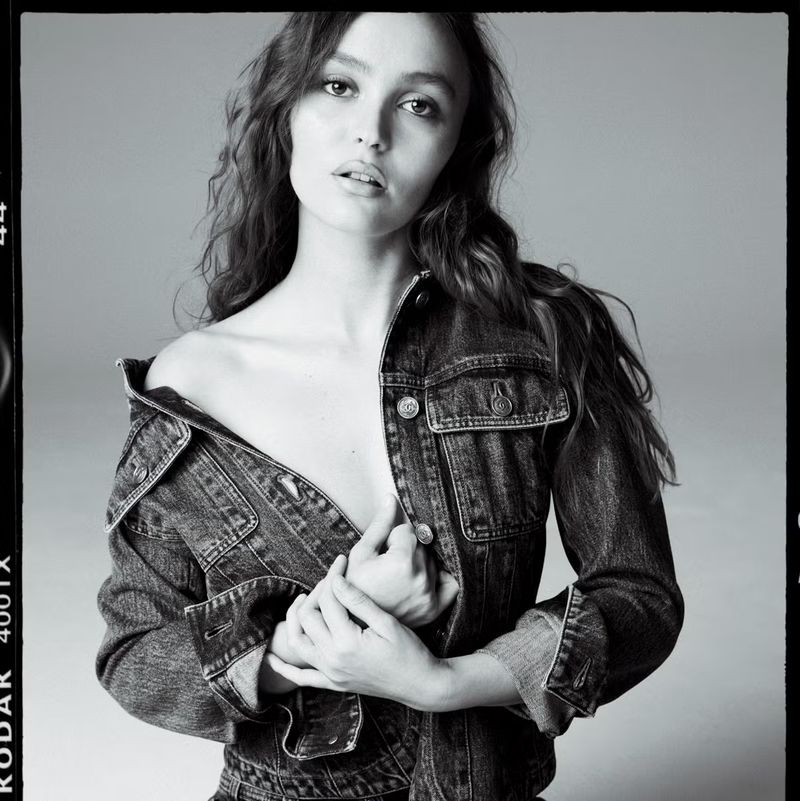 Monochrome portrait of Lily-Rose Depp wearing an off-the-shoulder denim jacket against a neutral grey studio background.